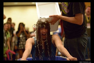 A woman is seen yelling while a bucket of ice water is being poured over her head. She’s in the middle of the room surrounded by dozens of people in the background, out of focus, seemingly cheering her on.