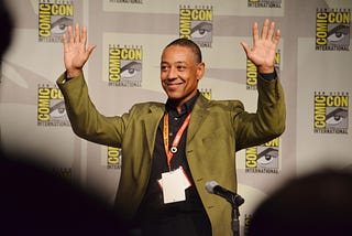 Giancarlo Esposito waves to a crowd with both hands. He’s wearing a green jacket and standing in front of a Comic-Con backdrop.