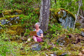 A woman sits with her back against a tree in westcoast forest. She has a computer in front of her
