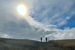Silhouette of two runners at the crest of a trail, set against a sky with sweeping clouds and a bright sun