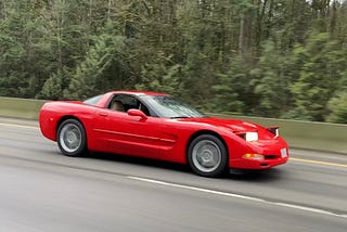 A torch red C5 Corvette driving through the forest at a speedy pace