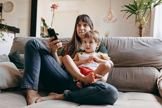 Image shows adult woman with small boy on her lap (possibly mother and son) sitting on a couch and looking ahead at what is presumably an off-camera television. Woman holds remote control.