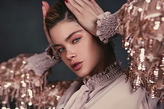 A young woman with glossy pink lips and shimmering eye makeup poses elegantly, wearing a blouse with sequin-embellished sleeves