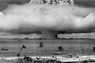 A black and white photo of a nuclear bomb going off in the sea with a palm treed beach in the foreground.