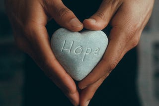 two hands holding a stone with the word “hope” etched on the surface. Photo by <a href=”https://unsplash.com/@ronkaowski?utm_content=creditCopyText&utm_medium=referral&utm_source=unsplash">Ronak Valobobhai</a> on <a href=”https://unsplash.com/photos/person-holding-gray-heart-shape-ornament-4qHWTuP_RLw?utm_content=creditCopyText&utm_medium=referral&utm_source=unsplash">Unsplash</a>