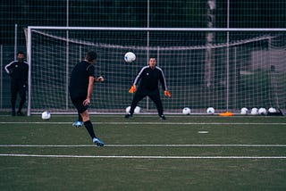 A soccer player in all black practicing shooting on a goalkeeper with orange gloves in goal.