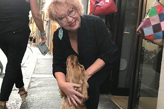 Author Carol Lennox in front of a store for hand painted purses. A cocker spaniel dog is looking up at her as she is smiling at the camera. Solo travel. Italy.