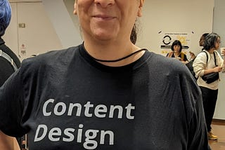 Mary Liebowitz, a pale woman with dark hair, smiles at the camera, wearing a black shirt with white text that reads “Content Design Manifestet http://thisiscontent.design”