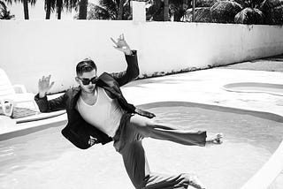 A black and white photo of a fully dressed young man falling sideways, mid-air, into an outdoor swimming pool. It looks hot because there are palm trees and he’s wearing shades.