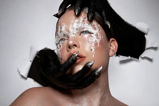 A woman with artistic white eye makeup poses against a white wall as two wolf-like hands clutch her chin and the top of her head respectively. She’s staring into the distance with a vacant expression.
