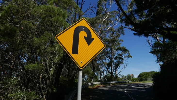 Yellow road sign with black curved arrow indicating turning around.
