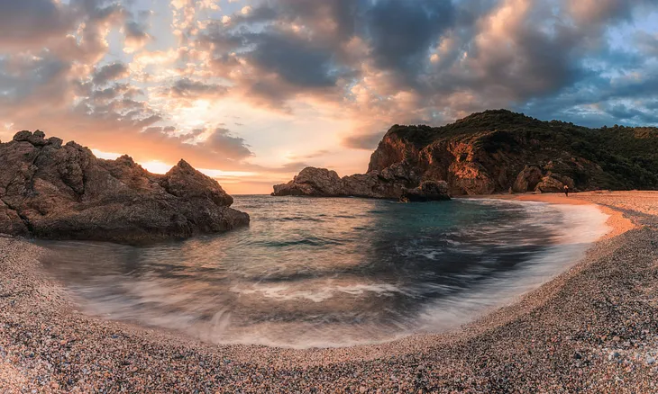A landscape photo of some water moving between two irregular shaped rocks, there is a pink-orange and blue tinge to the rocks, water and sky that makes one think of serenity.