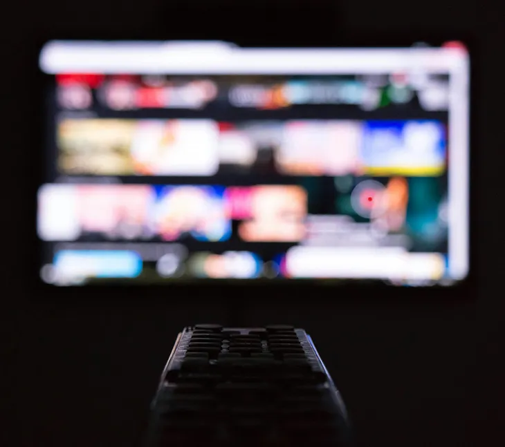 A blurred-out tv screen paused on a streaming platform. The focus is on a black remote pointed at the screen, which is enveloped in darkness.