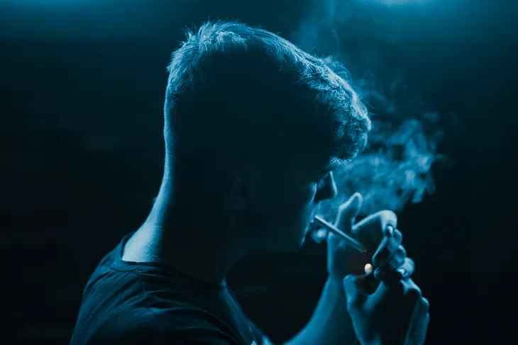 A male person in blue light lighting a cigarette.