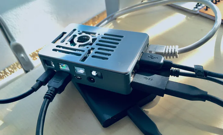 Photograph of a Raspberry Pi computer. It’s in a grey plastic housing and underneath is a SSD drive. Wires are sticking out from it on all sides.