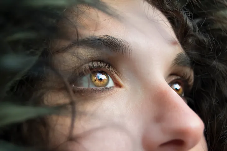 A woman with dark hair that is wind-blown around her face is looking upwards with gold brown flecked eyes that are full of anxiety. She is watching intently as if expecting something she is fearful of to happen.