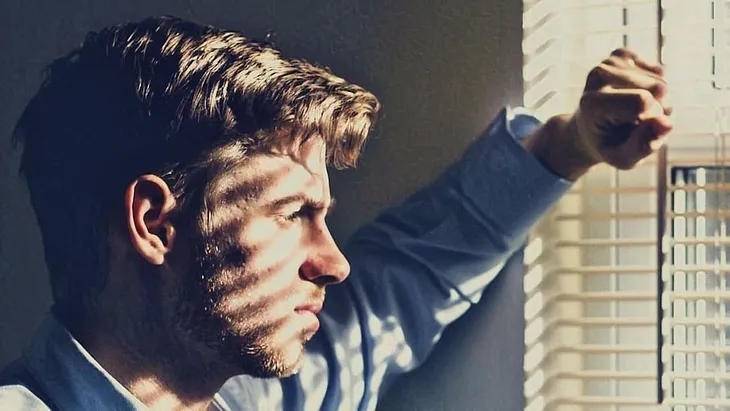 Pensive young man gazes out window. The blinds cast striped shadows on his face.