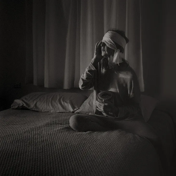 A black and white photo of a woman sitting on a bed with a bandage on her head