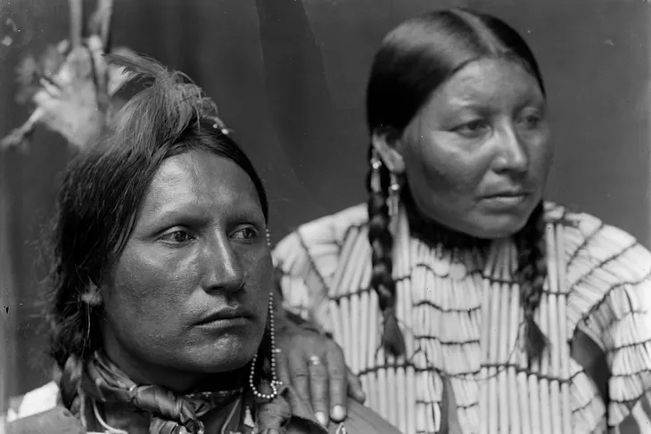 A Native American husband and wife, Mr. and Mrs. American Horse, look at something out of frame. She lays her hand on his shoulder.