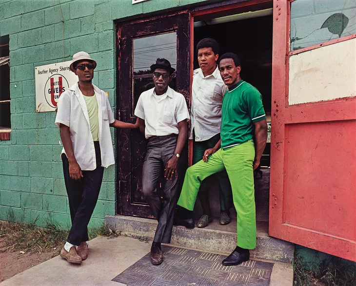 four Black men stand in an open doorway, green wall behind them, red door out to the side. Three of the men have white button ups and dark slacks, while one man has a green shirt and lime green pants. All look back towards the camera.