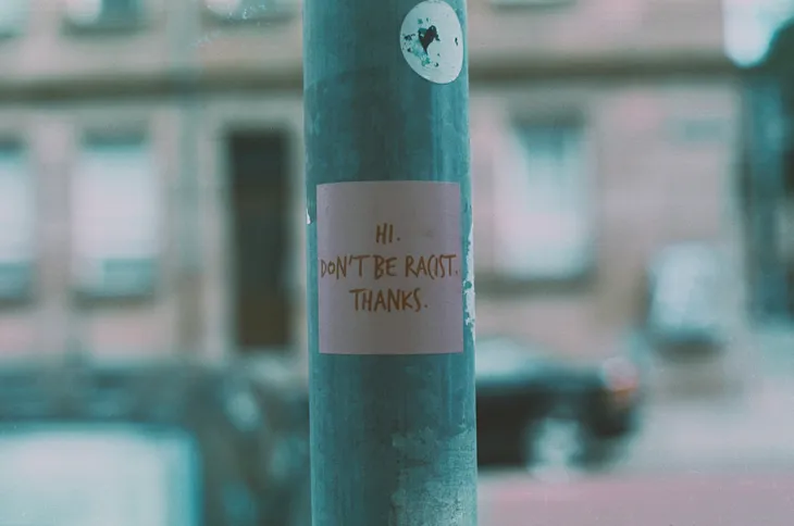 Sign posted on a pole with a blurred background. Sign reads “Hi. Don’t Be Racist. Thanks.”