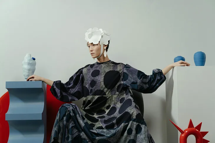Model wearing loose, spotted high-fashion dress and white cap with chin strap and sitting between blue and red modern art sculptures in front of a white background