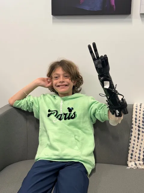 Photo of child wearing upper limb prosethetic. They are sitting on a couch giving the peace sign with the prosethetic and smiling.