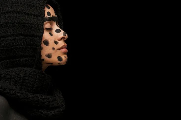 A serene woman with a thoughtful expression, her face artistically adorned with black paint spots resembling ellipses. She wears a black scarf, set against a stark black background, a contemplative mood that resonates with the topic of the poem ‘My Existence In Ellipses….’
