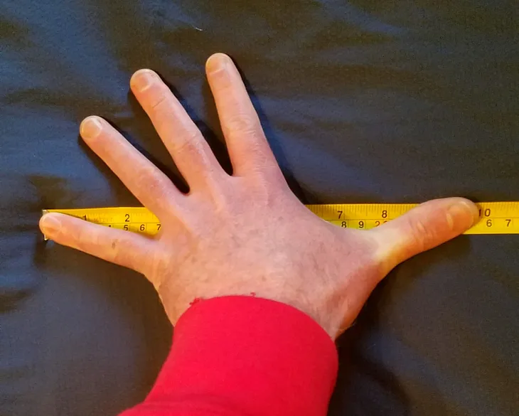 A photo of the author’s left hand with a tape measure showing a length of 9.75 inches from the tip of the pinky finger to the tip of the thumb.