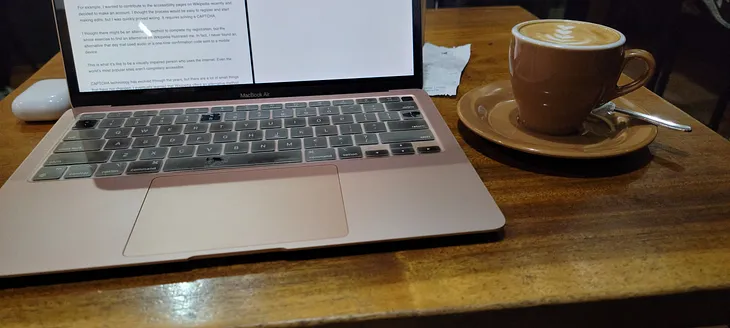 A Macbook Air, a latte and a Airpods case on a wooden table somewhere in Asia.