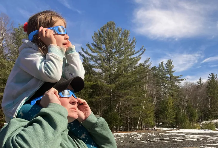 4-year-old on Mom’s shoulders looking skyward with eclipse glasses and expressions of awe during the total solar eclipse on April 8, 2024