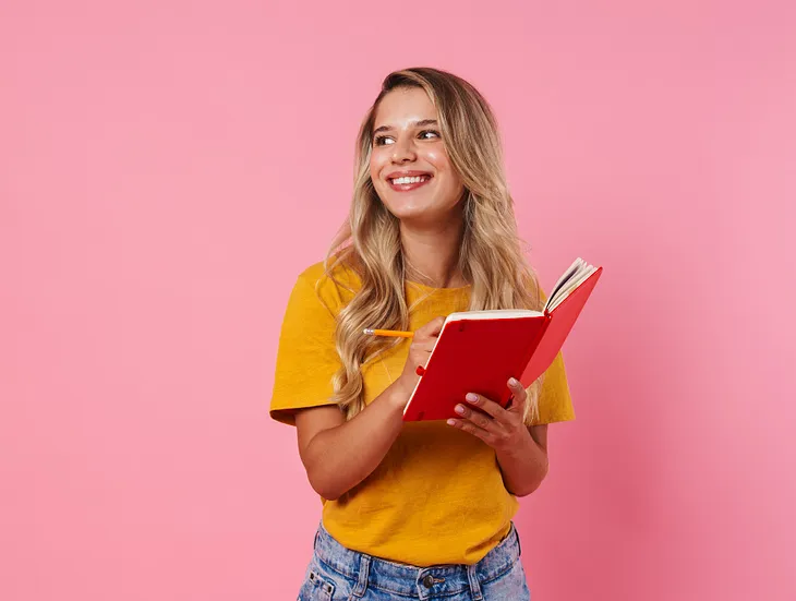A smiling blond woman writing in a red notebook on a pink backdrop