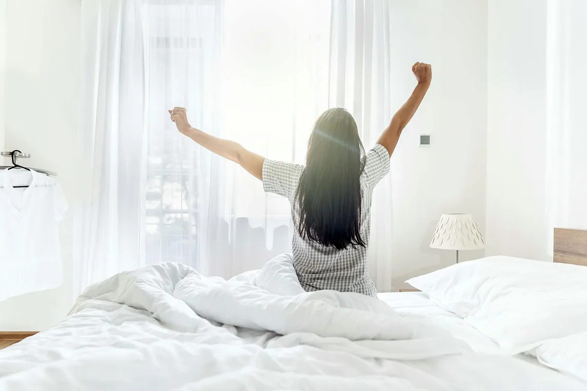 A woman sitting up in bed and stretching as the morning light streams in through a window
