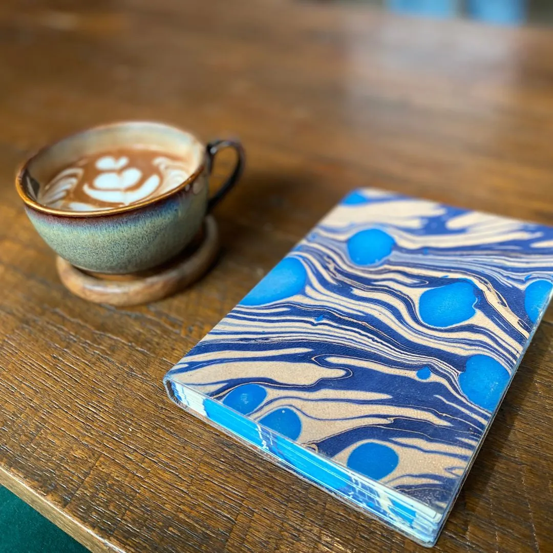 A marbled blue journal next to a cup of coffee with flower latte art on a wooden table