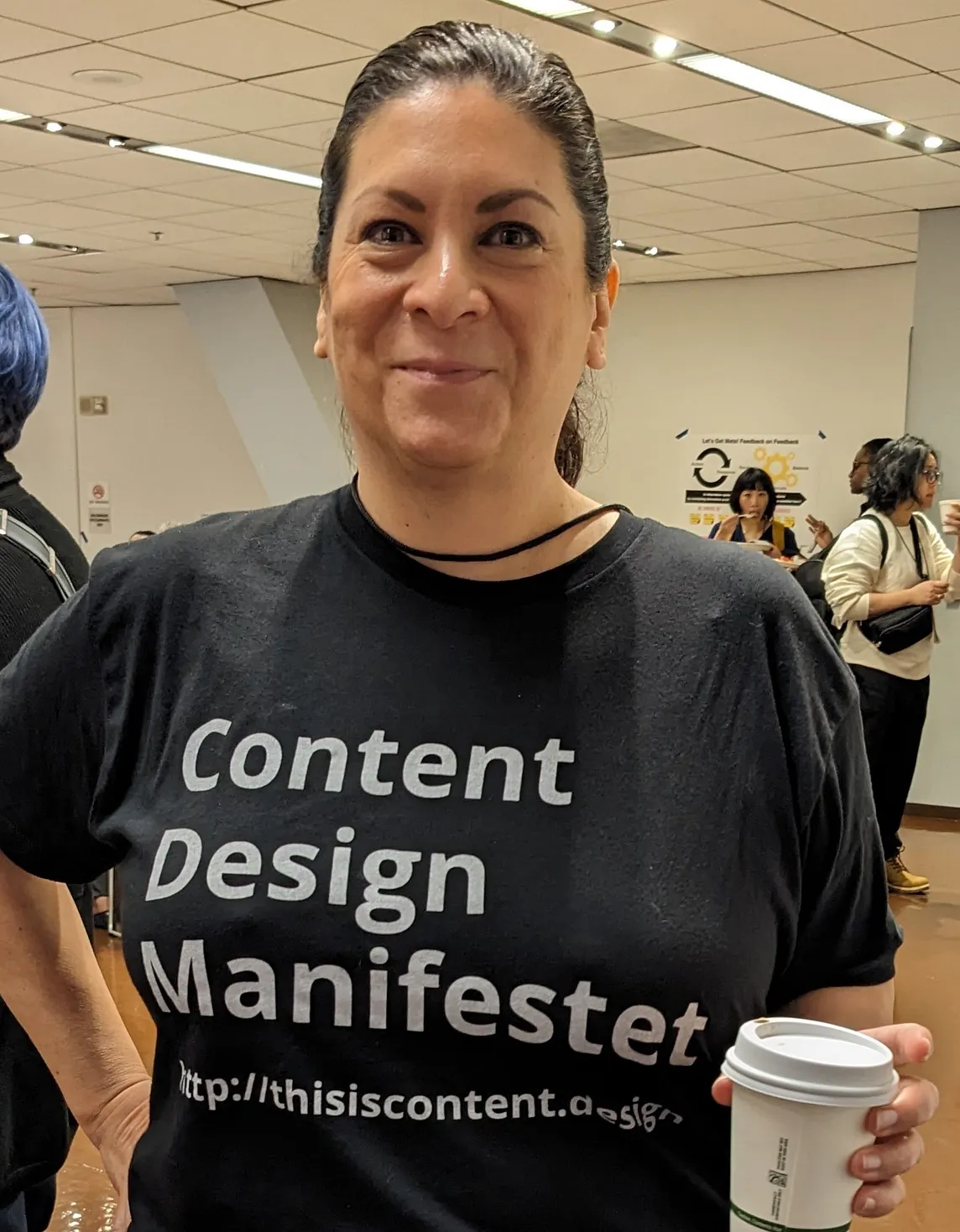 Mary Liebowitz, a pale woman with dark hair, smiles at the camera, wearing a black shirt with white text that reads “Content Design Manifestet http://thisiscontent.design”