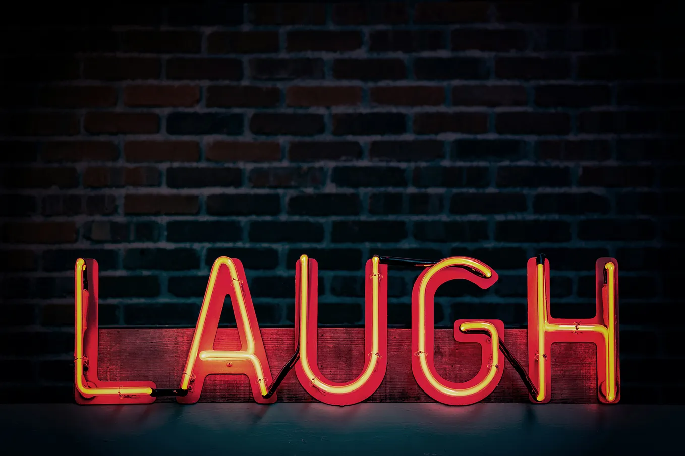 Coloured image in which a neon sign spells out the word ‘laugh’ in bright letters against a dark brick wall.