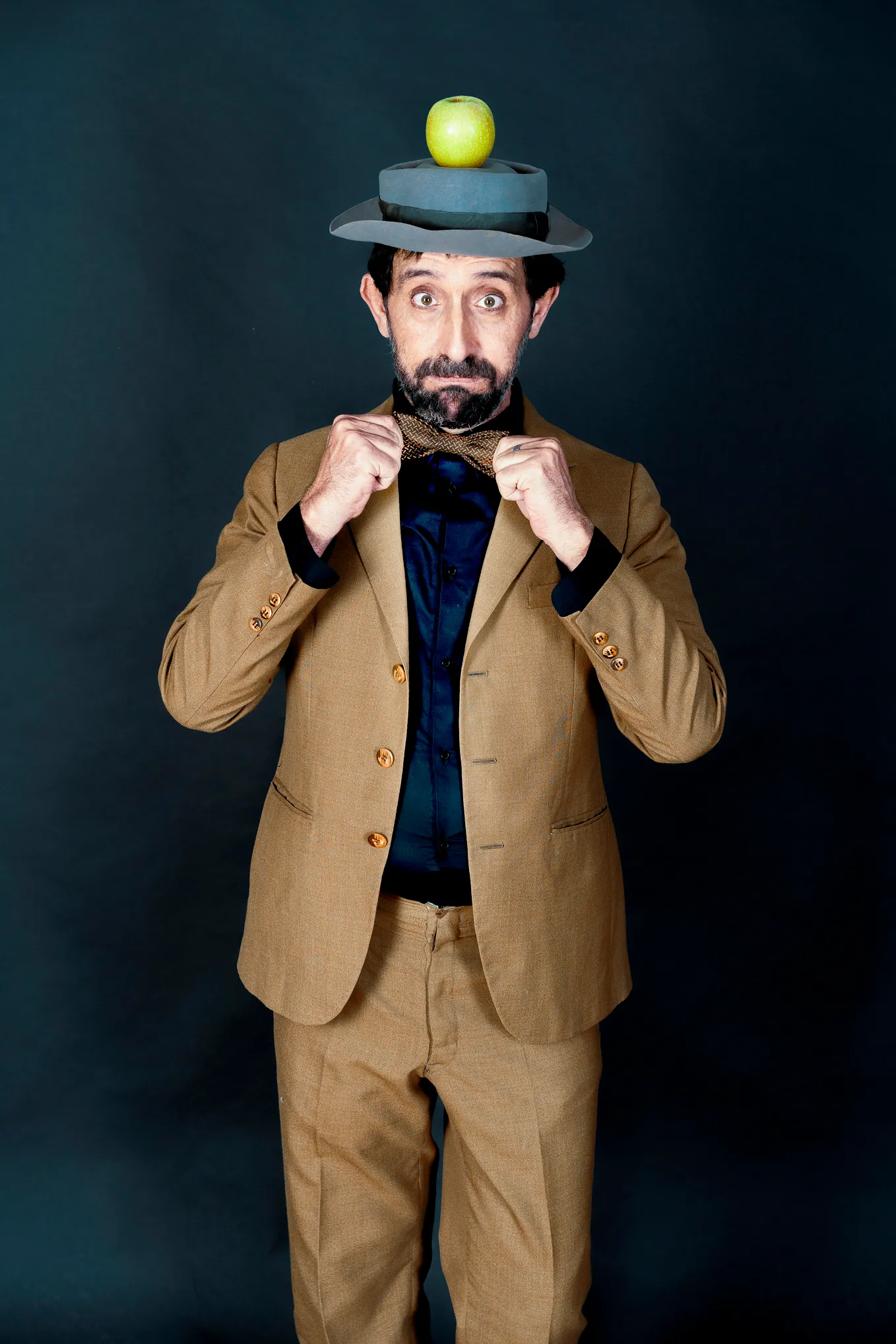 Coloured image shows a man standing against a dark background. He is wearing a brown suit and navy shirt. He has a felt hat on his head, and an apple sits on top of the hat. He has a dark beard and a perplexed expression. He is in the act of adjusting his bow tie.